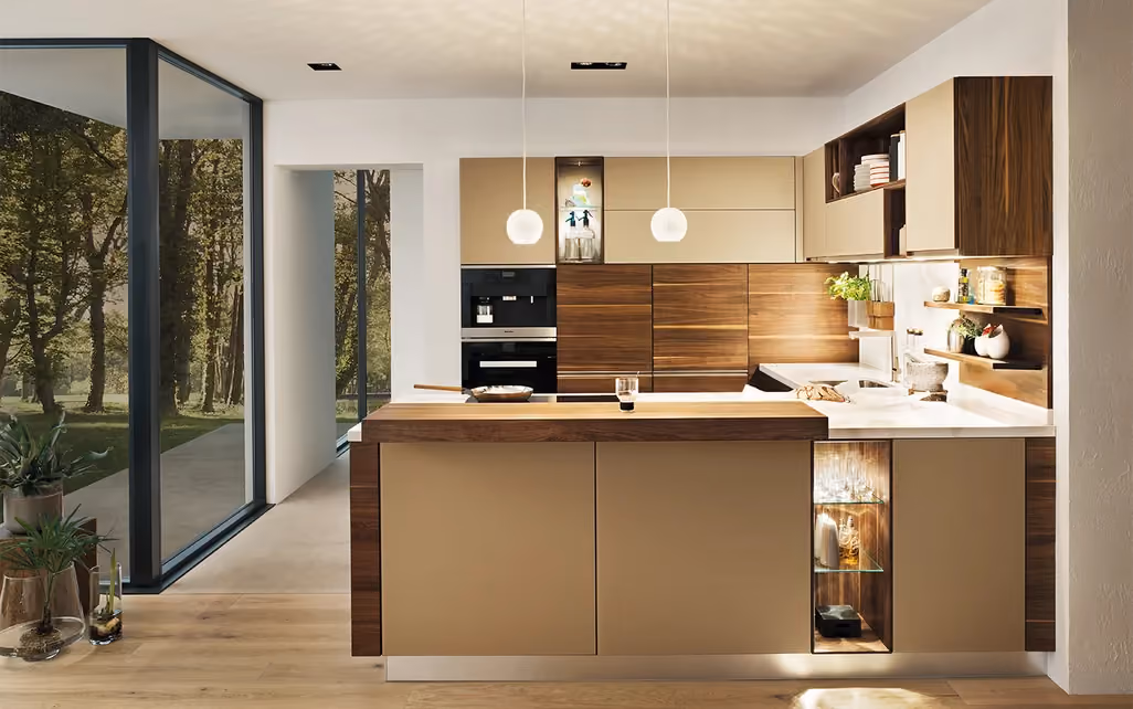 Modern kitchen with an island featuring beige and wood paneling, two hanging pendant lights, built-in appliances, and large floor-to-ceiling windows showing a green outdoor area.
