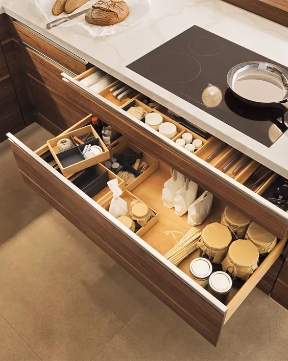 Open kitchen drawer with organized compartments holding various jars, bottles, utensils, and kitchen items below a countertop stove with a pan on it and bread on a cutting board.