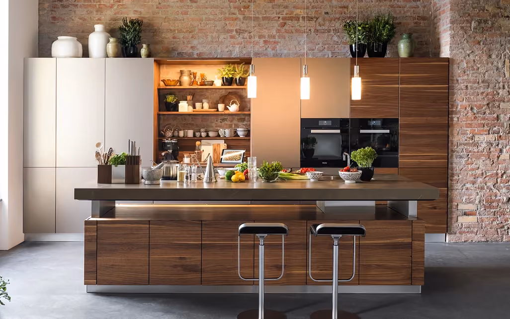Modern kitchen with a large island featuring dark wooden cabinets, two black stools, pendant lights, and a variety of plants and kitchen items, set against exposed brick walls with built-in wood shelves and ovens.
