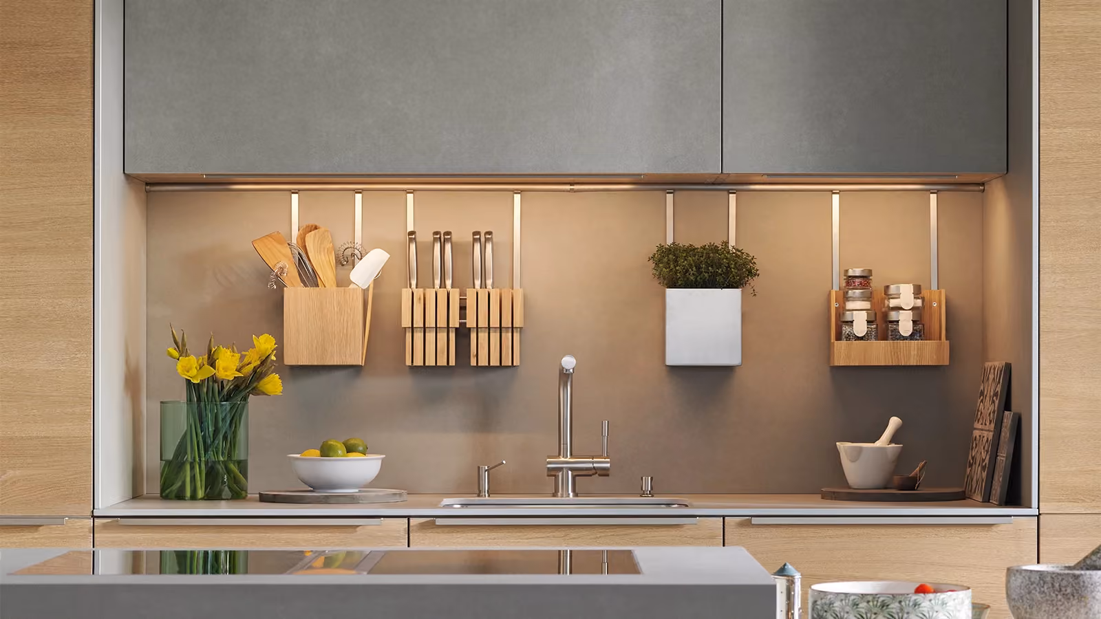 Modern kitchen backsplash with a mounted rail system holding a wooden utensil holder, knife blocks, a planter with greenery, and a spice rack, above a sink with a stainless steel faucet, surrounded by light wood cabinetry and under-cabinet lighting.
