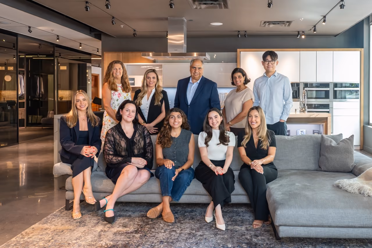 Group of nine diverse people posing in a modern office lounge area with a grey couch and kitchen in background.