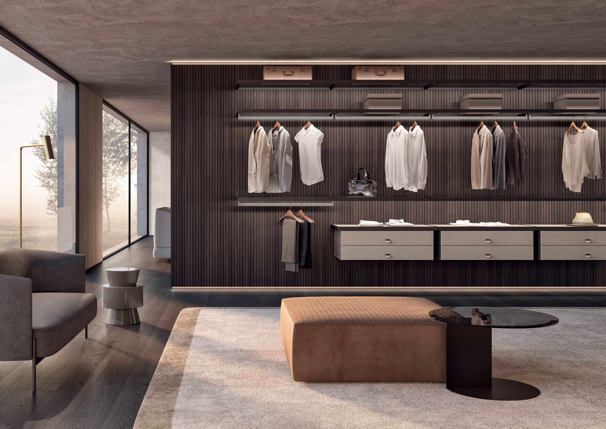 Modern walk-in closet with dark wood panel wall and hanging clothes including shirts, jackets, and pants, two suspended drawer units, and a brown ottoman with a small round black coffee table on a light gray rug. Large window with a view of trees and seating area with two gray armchairs and a small side table are visible to the left.