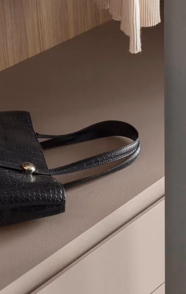 Black textured handbag with gold clasp resting on a taupe desk near a beige sweater and wood panel.