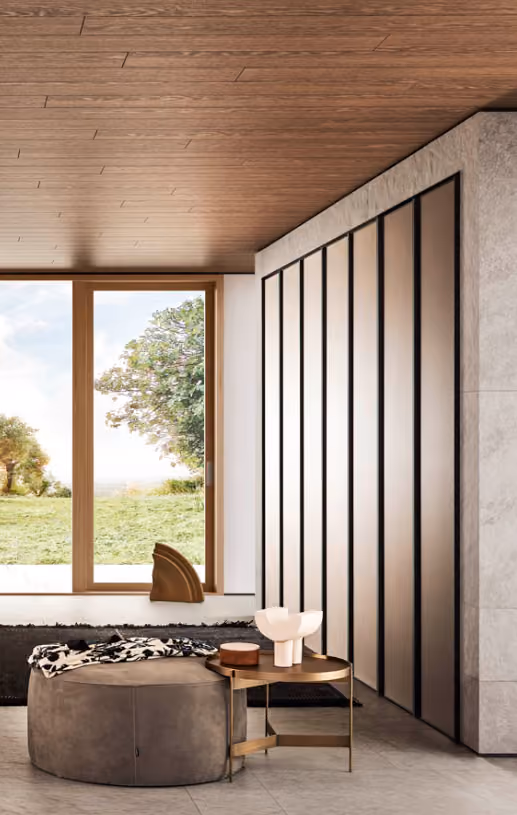 Modern room with wooden ceiling, large window overlooking grass and trees, and a round ottoman with a side table.