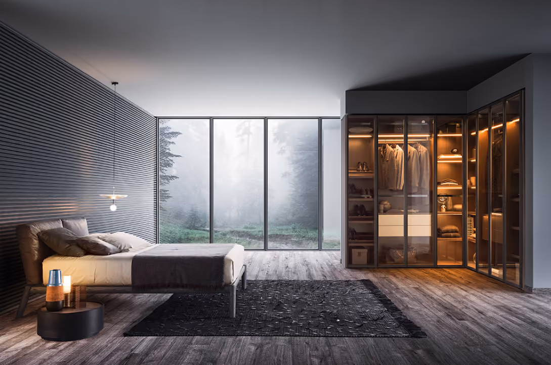 Modern bedroom with a low bed against a dark textured wall, bedside table with a lamp and cup, large floor-to-ceiling windows showing a foggy forest outside, and an illuminated walk-in closet with clothes and shelves.