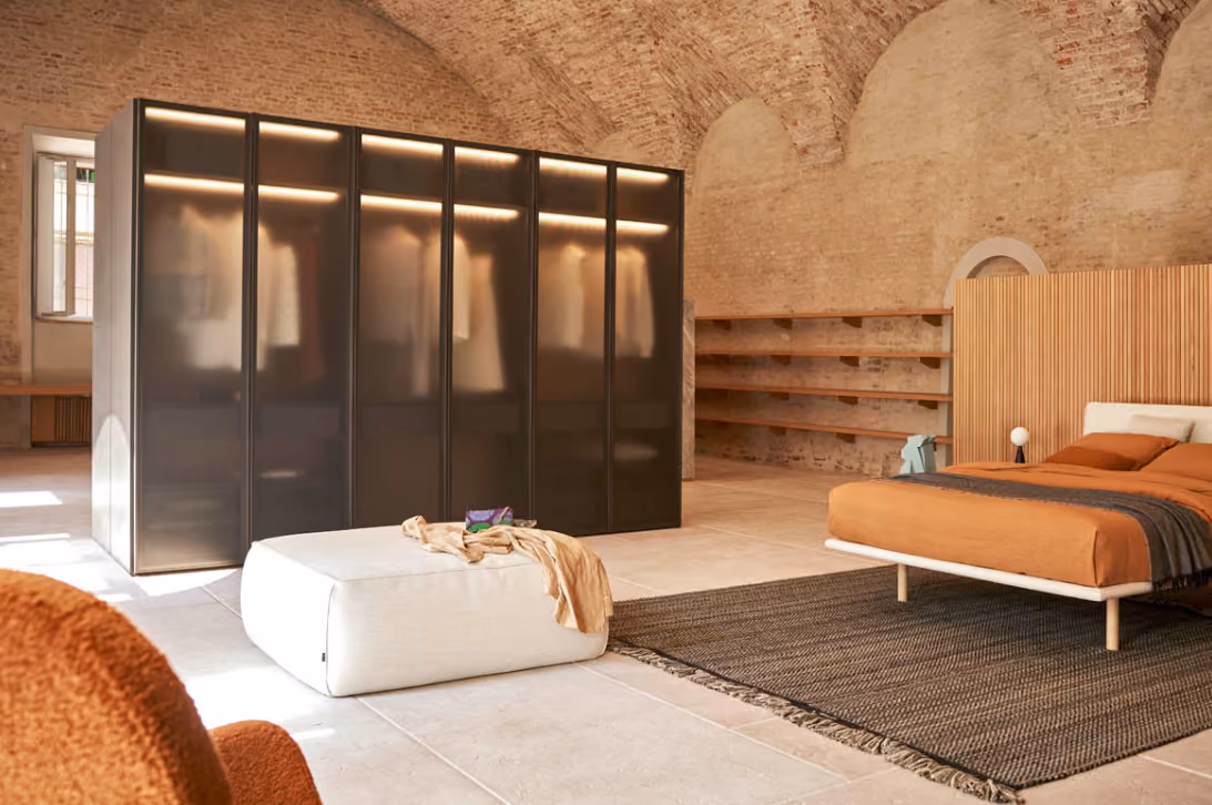 Modern bedroom with curved brick ceiling, featuring a burnt orange bed on a dark rug, a large frosted glass wardrobe, white ottoman, and wooden shelves.