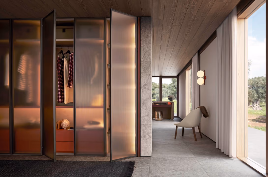 Modern interior hallway with frosted glass closet doors partially open showing clothes inside, a light-colored armchair against a wall with round wall lamps, large windows with sheer curtains, and a view of trees outside.