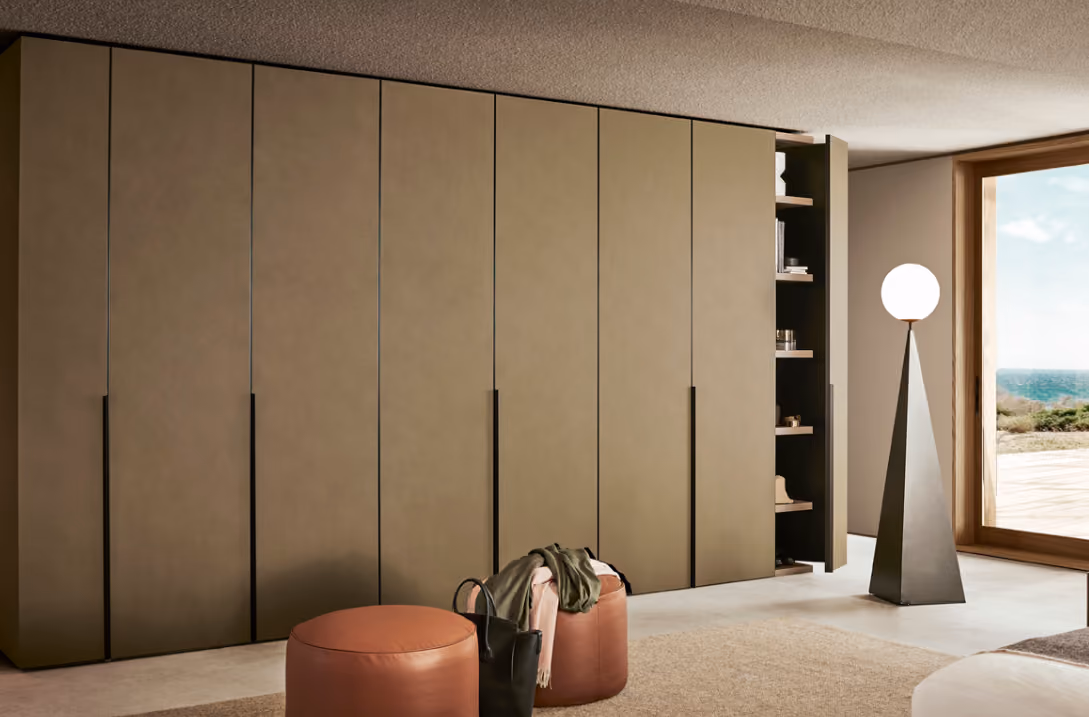 Modern room featuring a long wall of closed beige cabinets with one cabinet door open revealing shelves with assorted items, beside a large window overlooking a landscape, and two round brown ottomans with a black bag and clothing on top, along with a tall floor lamp with a spherical white shade.