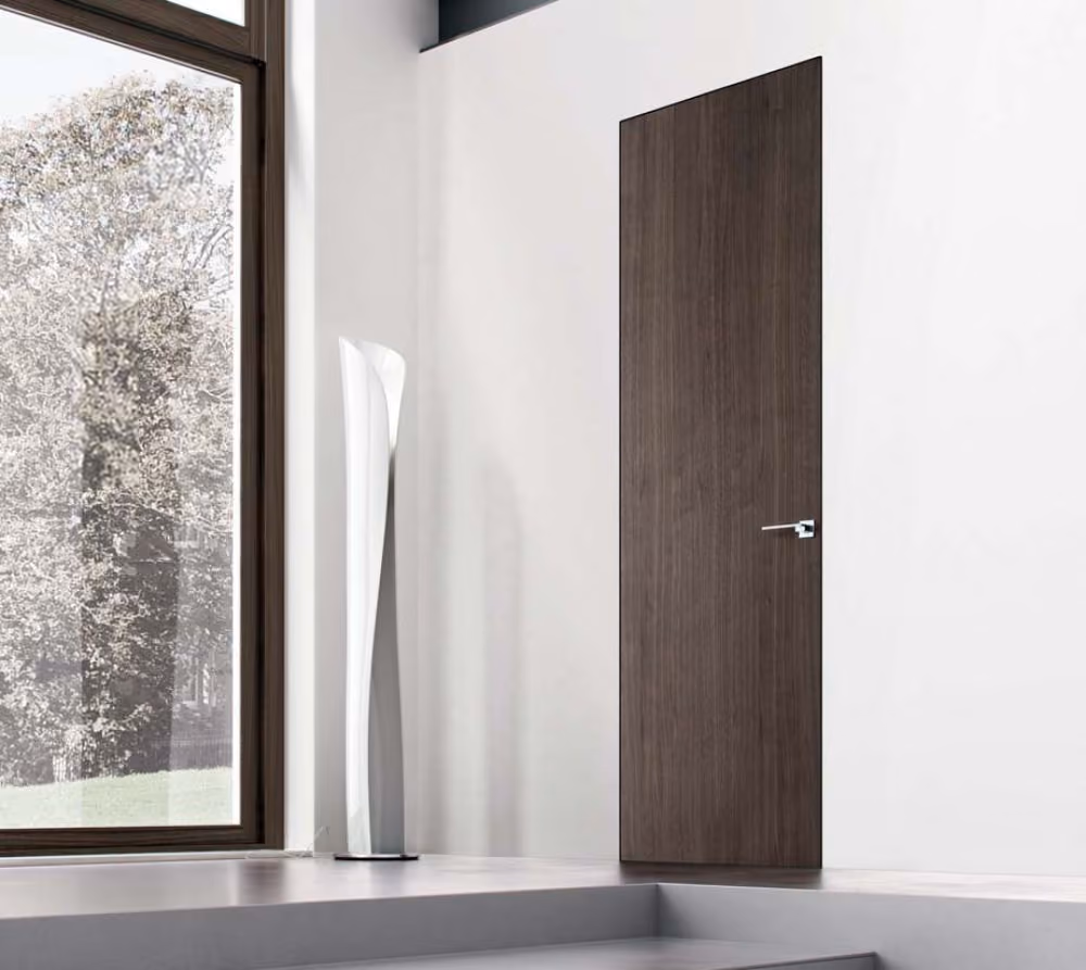Modern interior with a tall dark wood door featuring a sleek metal handle, a white minimalist sculpture beside it, and a large window showing trees outside.