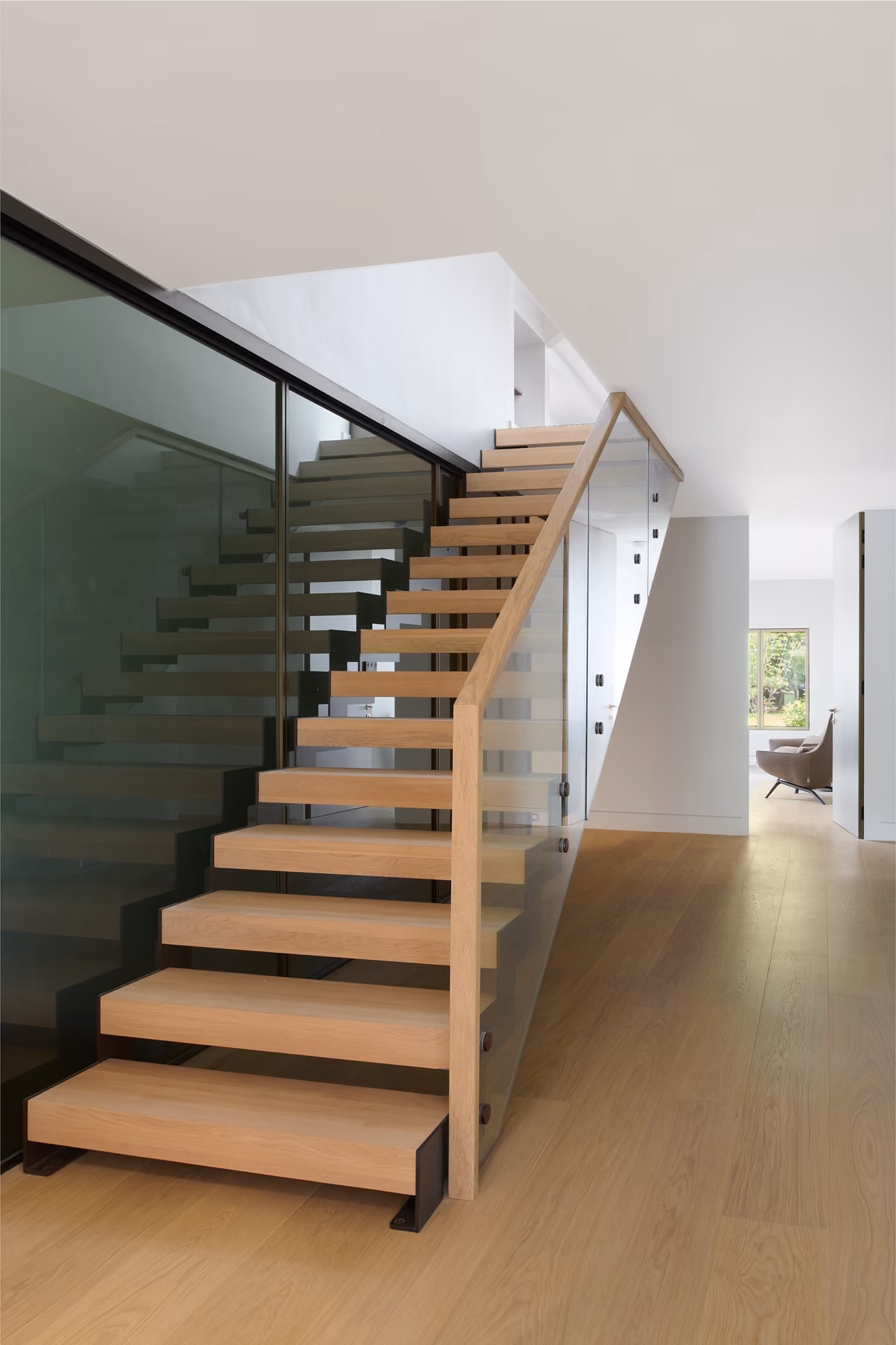 Modern interior with wooden floating stairs featuring glass side panels and light wood flooring, leading to an upper floor in a minimalist home.