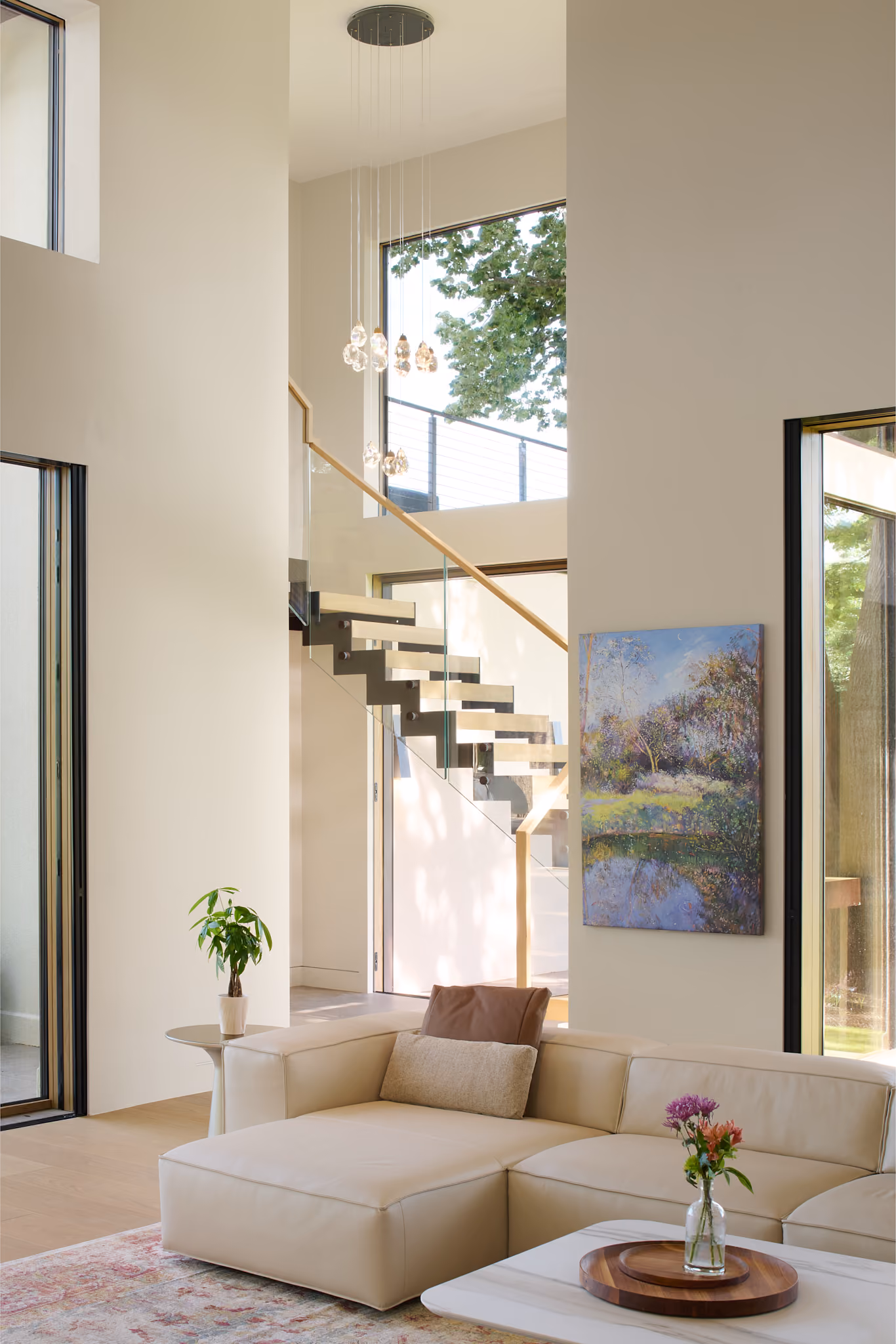 Modern bright living room with beige sectional sofa adorned with two pillows, a small plant on a side table, a coffee table with a flower vase, large windows, a nature painting on the wall, and a floating staircase with glass railing leading up beside a tall window showing green trees outside.