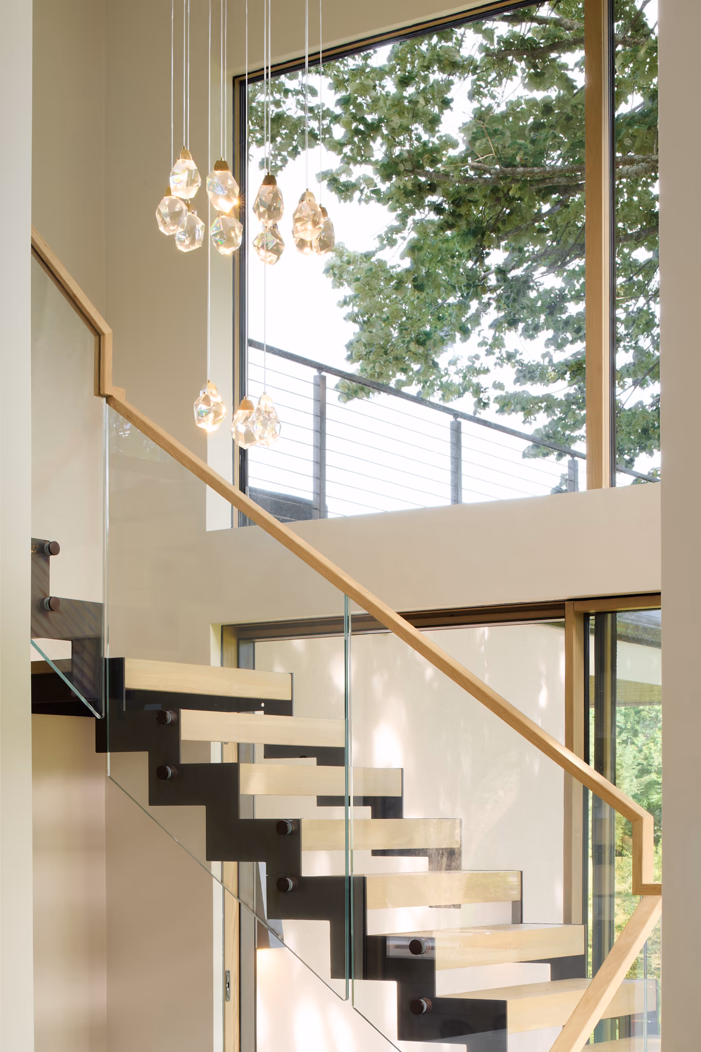 Modern staircase with floating wooden steps and glass railing next to large windows showing green tree branches outside, lit by hanging cluster of crystal pendant lights.