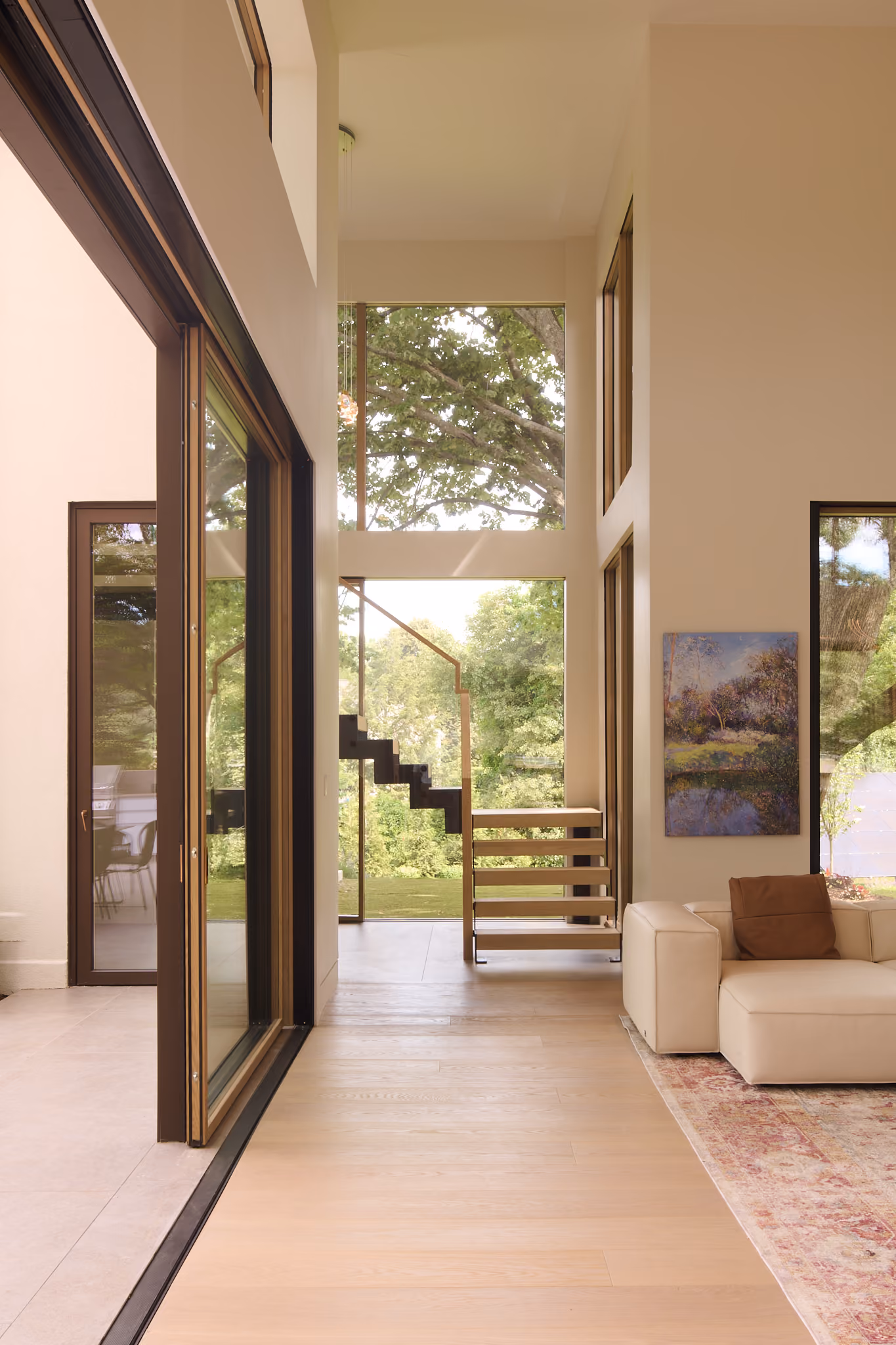 Modern interior with tall windows showing green trees outside, floating wooden stairs, beige L-shaped sofa with brown pillow, and a landscape painting on the wall.