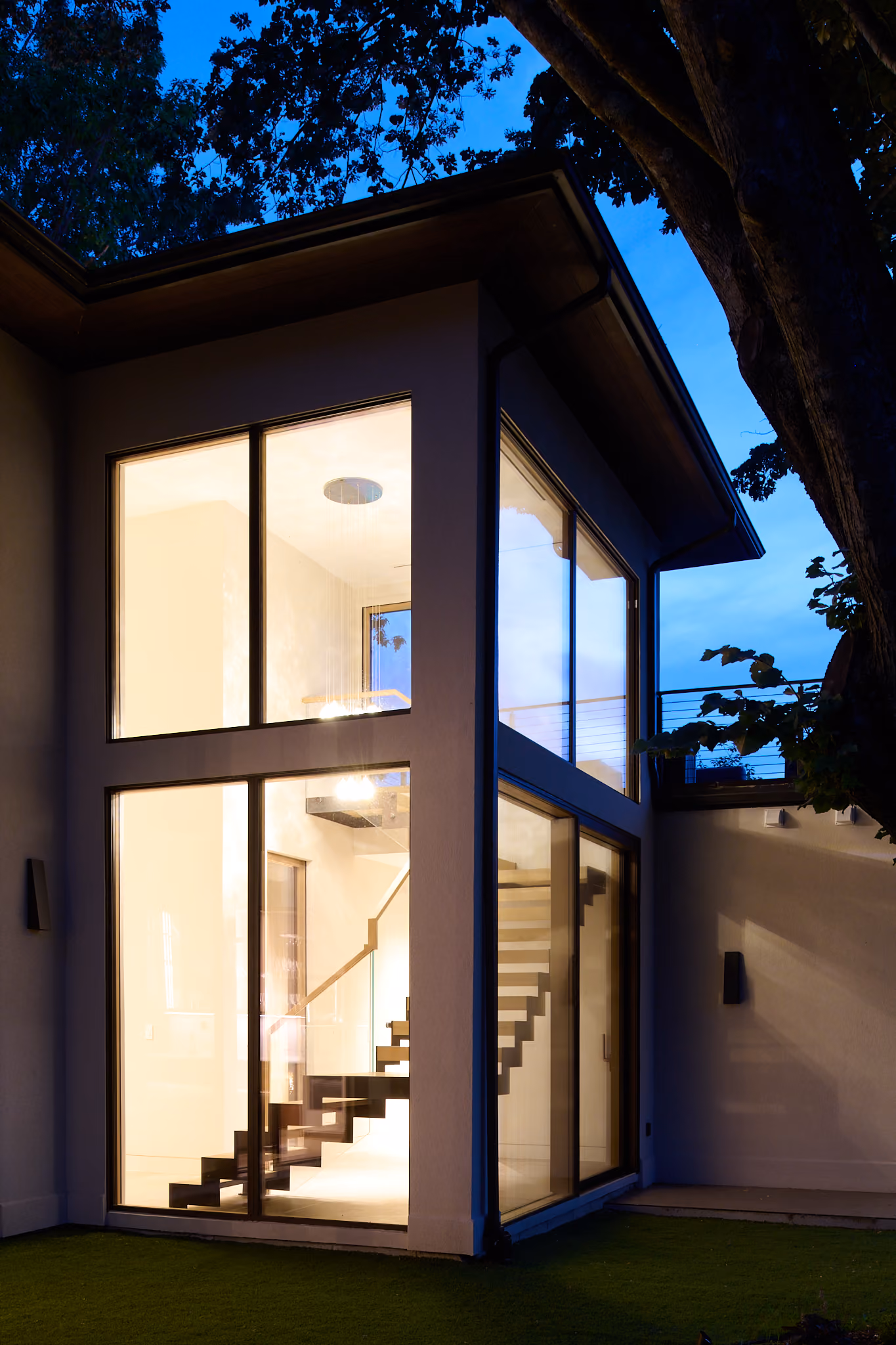 Exterior view of a modern two-story house corner at dusk with large illuminated glass windows showing an interior staircase and warm lighting.