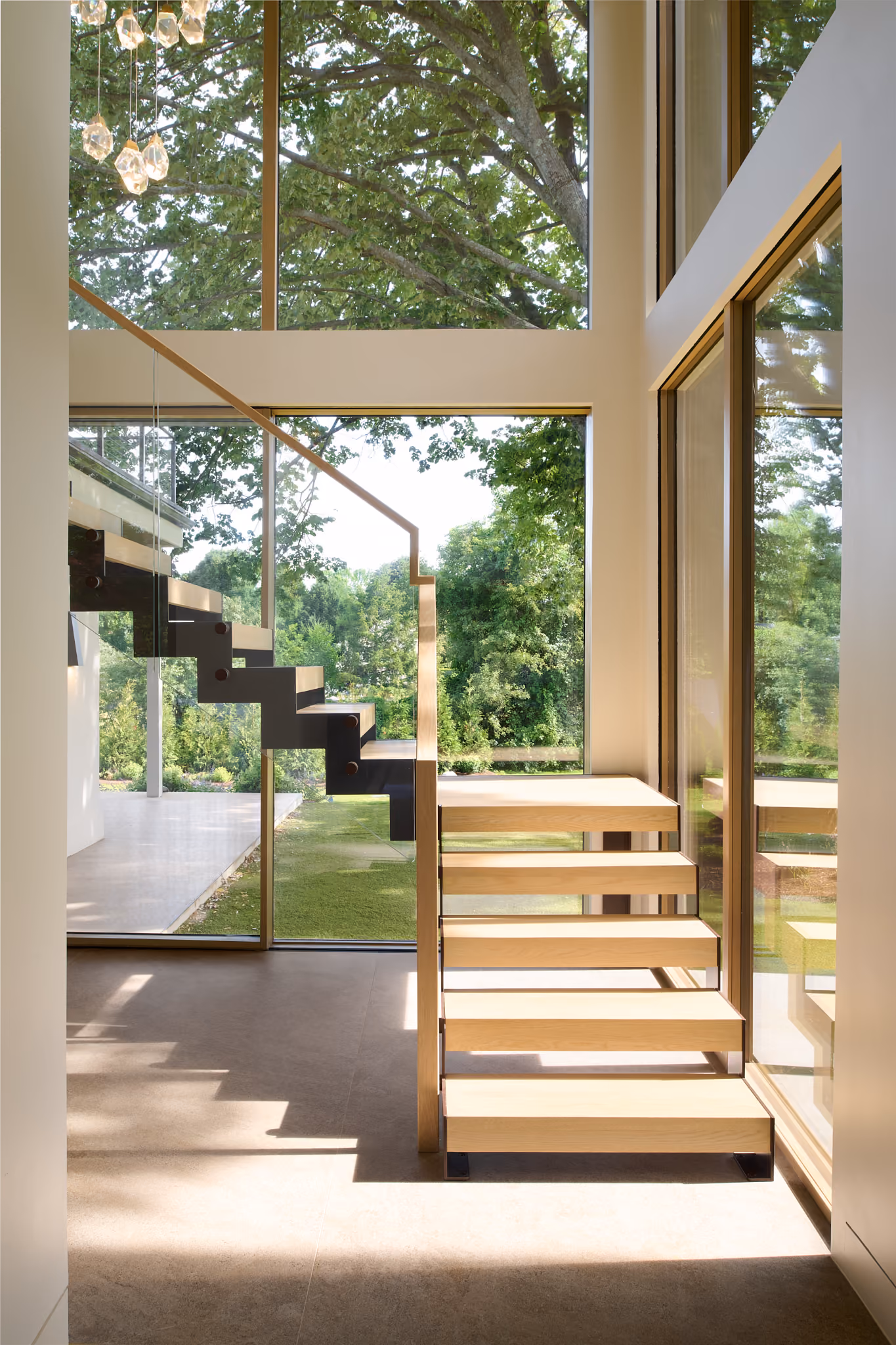Modern wooden staircase with glass railing inside a house with large windows showing green trees outside.