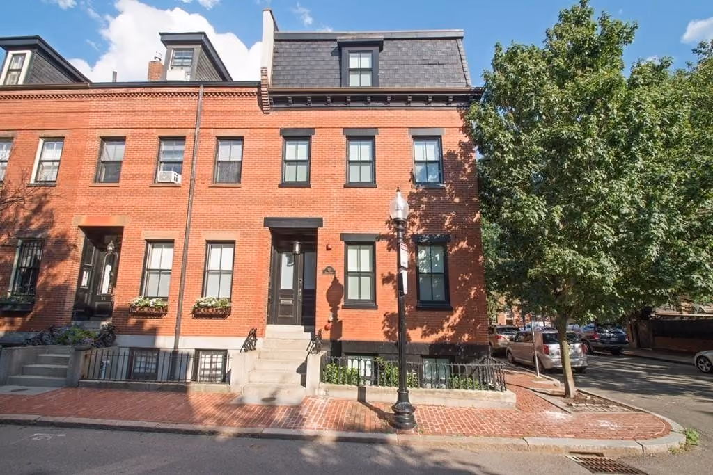 Three-story brick townhouse with black window frames, a black front door, and a mansard roof, situated on a corner with a streetlamp and tree nearby under a blue sky with clouds.