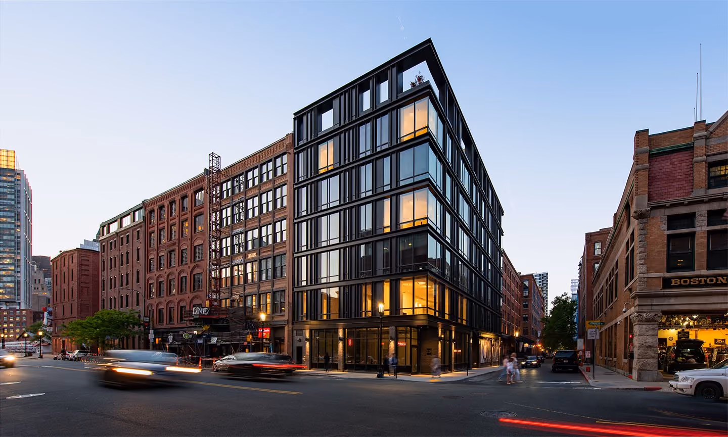 Modern glass and steel building at intersection in an urban area during early evening with older brick buildings on either side and light trails from moving cars.