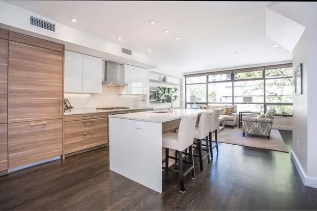 Modern open kitchen and living area with dark hardwood floors, white island with four beige bar stools, wood-finish cabinets, stainless steel range hood, large windows, and seating area with patterned armchairs.
