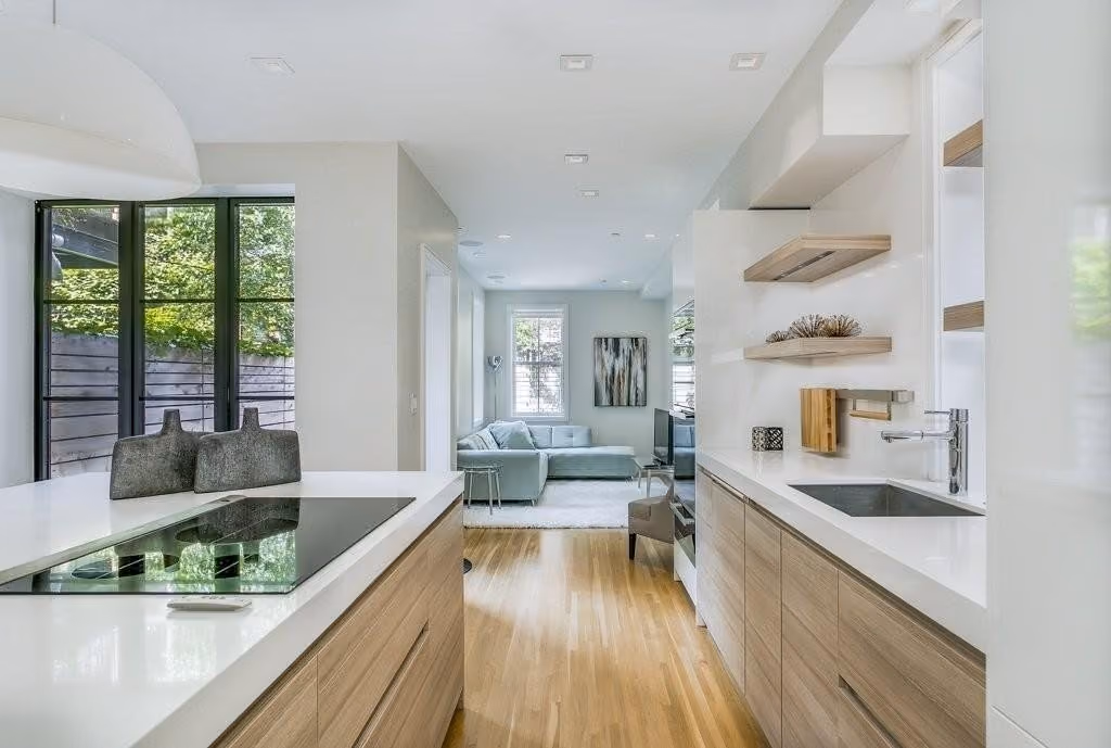 Modern kitchen with wooden lower cabinets, white countertops, and a built-in stovetop island, leading to a living area with a gray sectional sofa and abstract wall art.