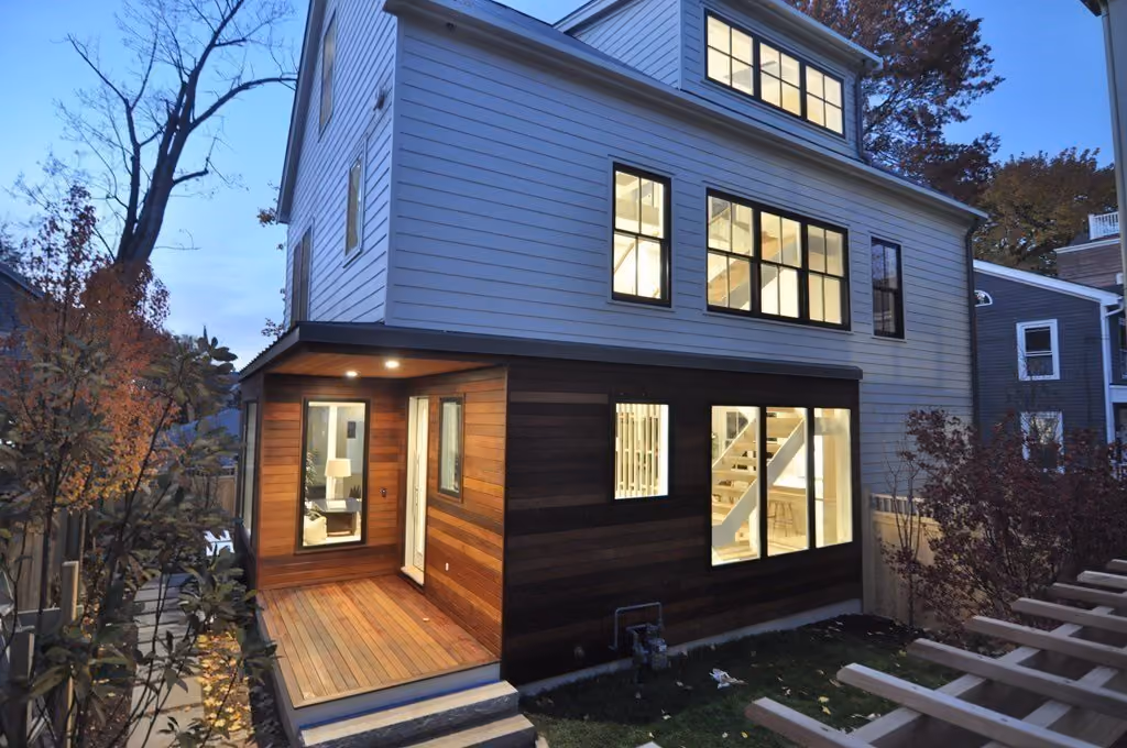 Modern two-story house exterior at dusk with white siding and a wooden porch area, large lit windows revealing stairs and interior rooms.