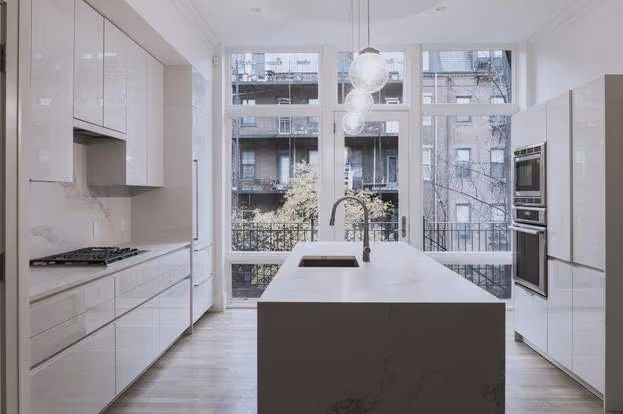 Modern kitchen with white cabinetry, a central island with a sink and faucet, pendant lights above, and large floor-to-ceiling windows showing a view of buildings outside.