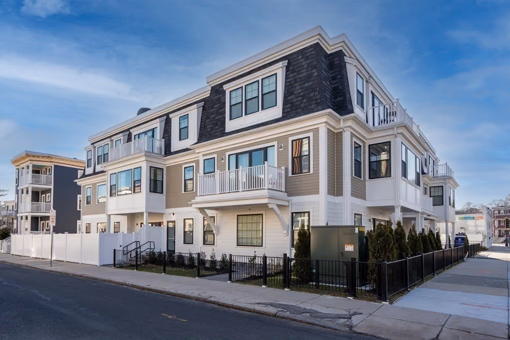 Three-story residential building with beige and white exterior, black roof shingles, balconies, and black metal fences along the sidewalk under a blue sky.