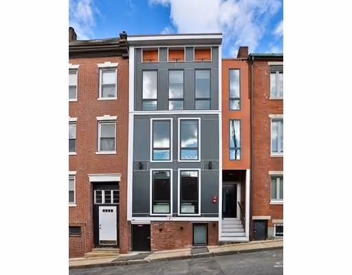 A narrow modern three-story townhouse with a gray facade and large rectangular windows, flanked by traditional red brick buildings on both sides, situated on a slightly sloped street under a partly cloudy blue sky.