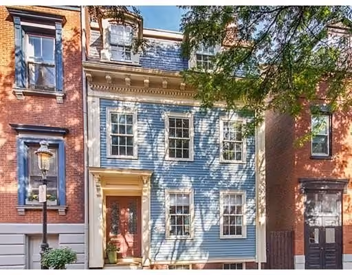 A classic blue wooden townhouse with white trim, featuring six windows and a red front door, flanked by taller brick buildings under a clear sky with tree shadows on the facade.