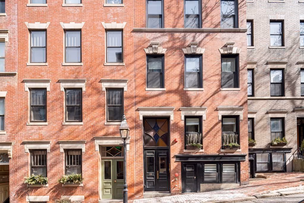 Street view of three adjacent multi-story brick buildings with distinct front doors, several windows with stone lintels, and window boxes with greenery, under daylight with shadow patterns on the façades.