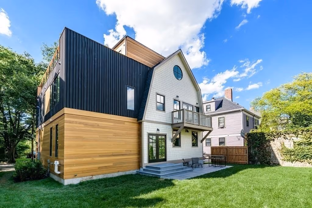 Modern three-story house with a mix of black vertical siding, natural wood paneling, and white shingle siding, featuring large windows, a balcony, and a backyard patio with outdoor furniture on green grass under a blue sky with scattered clouds.