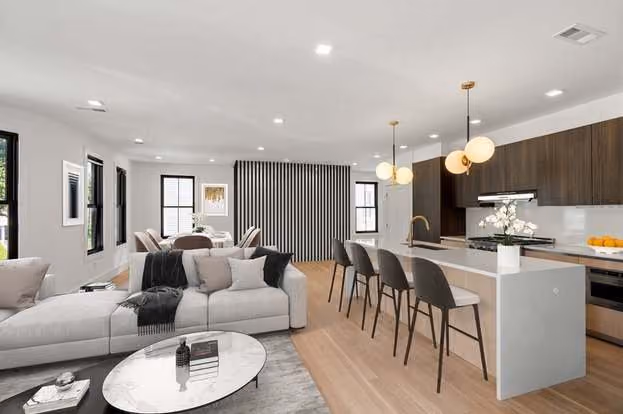 Modern open-plan living space with a light gray sectional sofa and pillows, round marble coffee table on a rug, adjacent dining area with table and chairs, and a kitchen with a white island, four high chairs, hanging globe pendant lights, and dark wood cabinets.