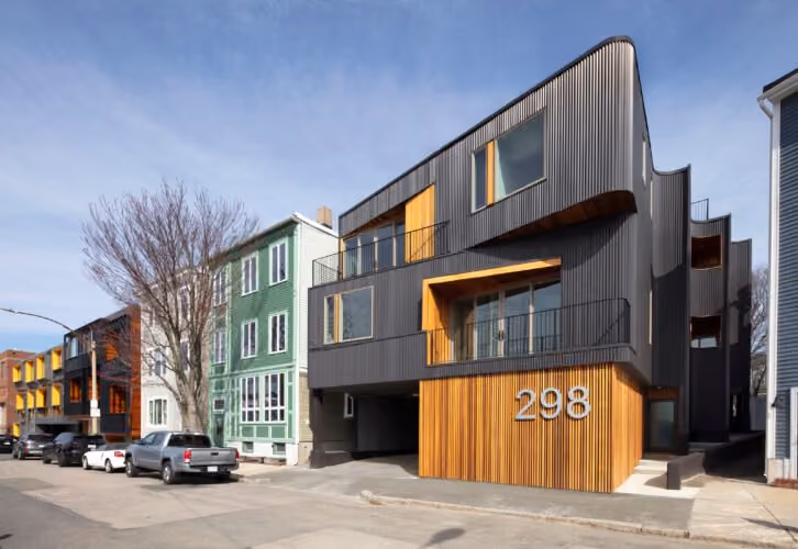 Contemporary three-story residential building with dark vertical siding and wooden accents, large windows, and the number 298 displayed on the front, situated on a street with parked cars and neighboring houses.