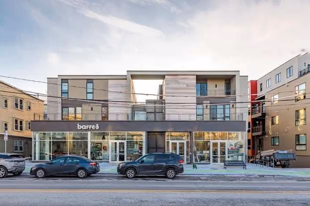 Modern two-story commercial building with large glass windows on the ground floor and residential units above, featuring a barre3 fitness studio, parked cars, and neighboring residential buildings.