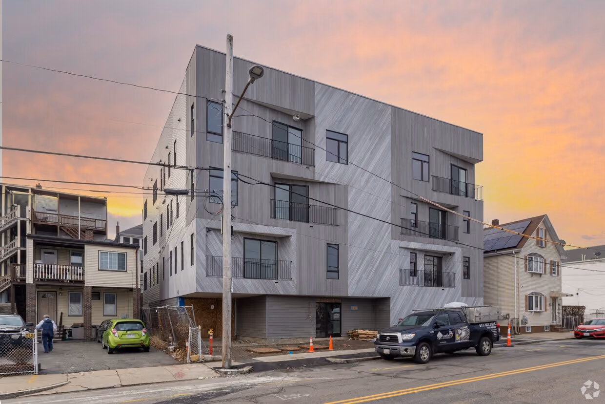Modern four-story apartment building with various-sized windows and balconies, situated on a city street at sunset, with cars parked along the curb and adjacent older residential homes.