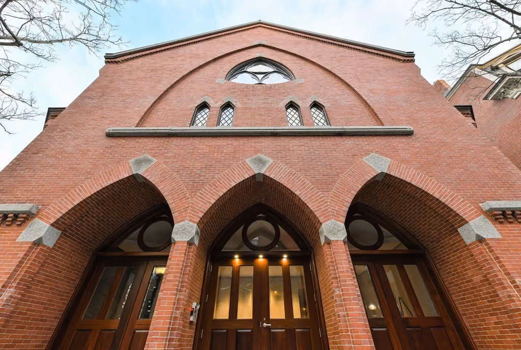 Upward view of a red brick building facade featuring three pointed arched doorways with wooden double doors beneath arched windows and decorative stone accents in gothic architectural style.