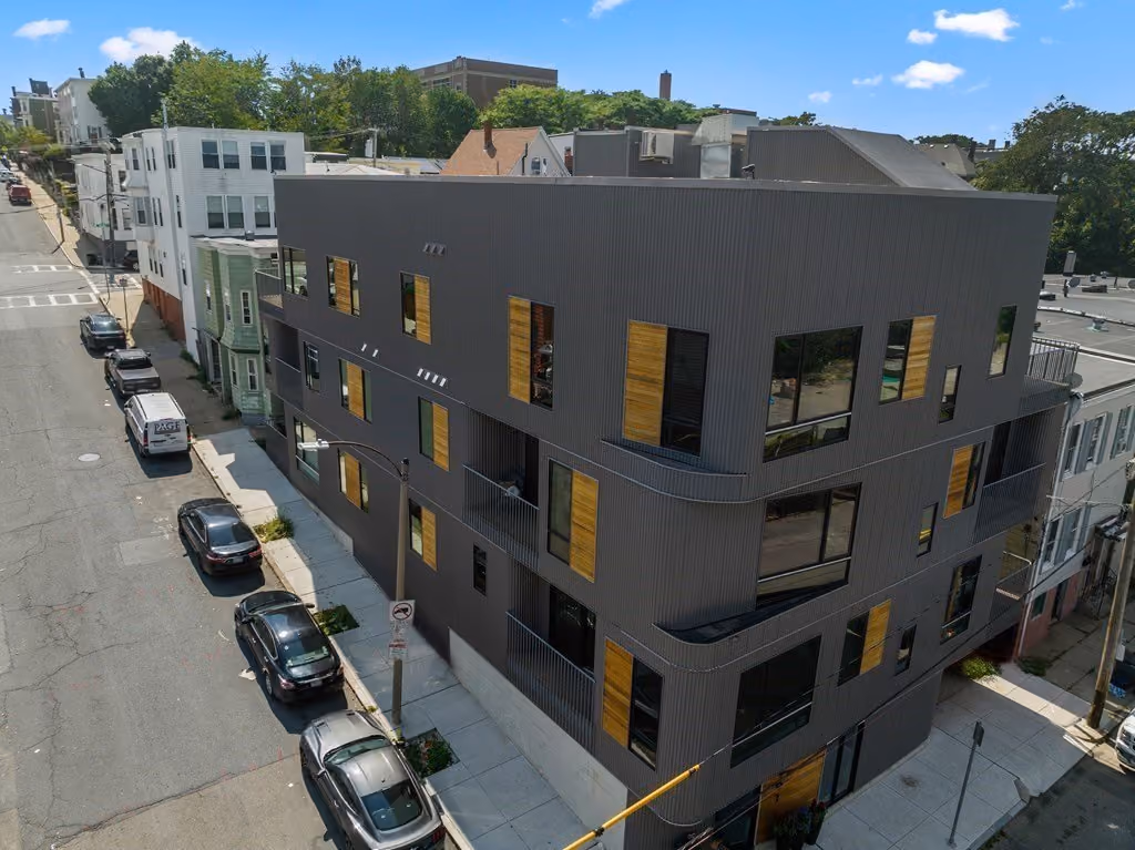 Modern three-story dark gray building with vertical ridges and wooden window accents on a city street corner with parked cars and adjacent older buildings.