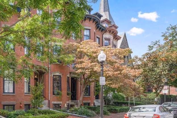 Street view of red-brick townhouses with ornate architectural details, trees with green and orange leaves, a black lamppost, and parked cars under a blue sky with scattered clouds.