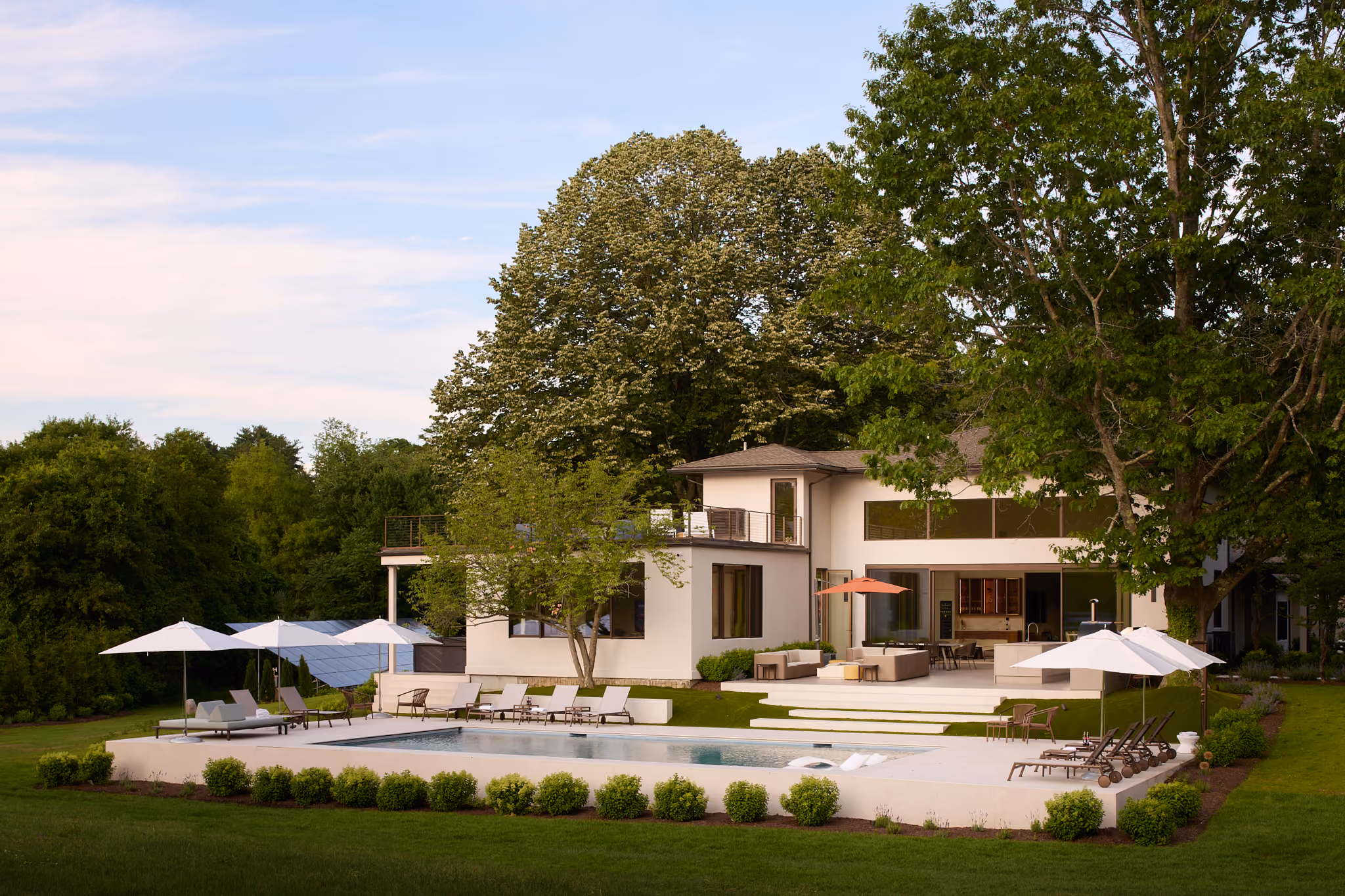 Modern two-story white house with large windows, a spacious patio, outdoor seating, umbrellas, and a rectangular swimming pool surrounded by green shrubs and lawn, set against tall trees and a partly cloudy sky.