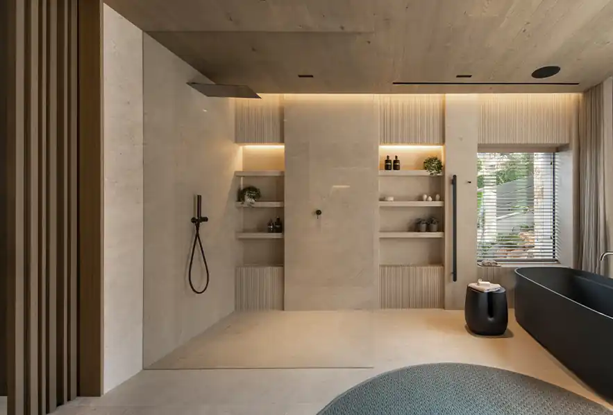 Modern bathroom with beige tiled walls and floor, featuring a glass-enclosed walk-in shower with black shower fixtures, built-in shelves with decorative items, a black freestanding bathtub near a window with blinds, and a small black stool with towels.