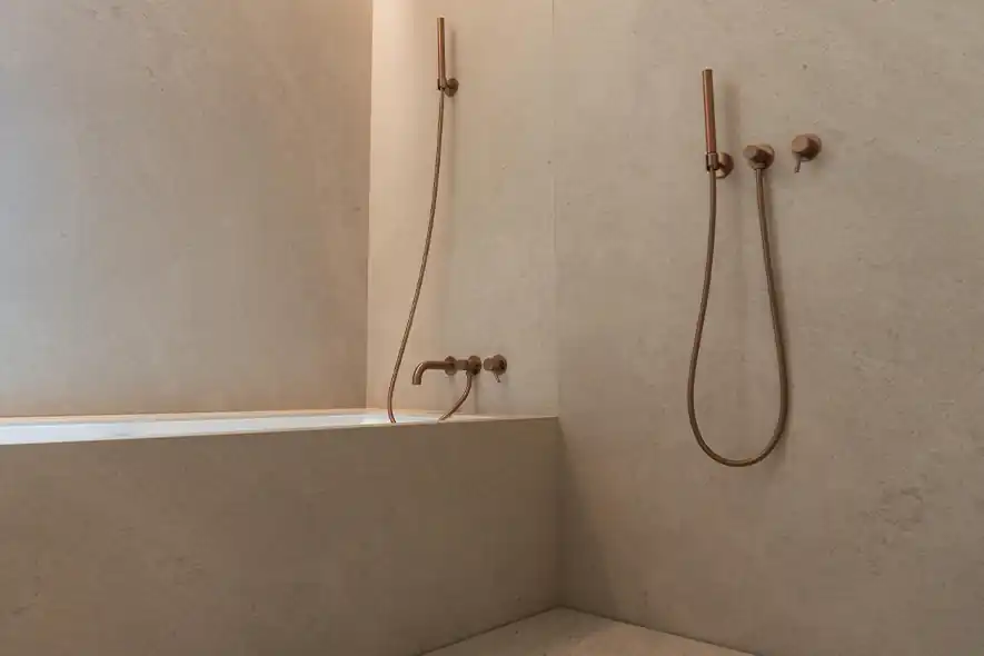 Minimalist bathroom corner with beige textured walls featuring a built-in rectangular bathtub and two wall-mounted handheld shower heads with metallic hoses and round controls.