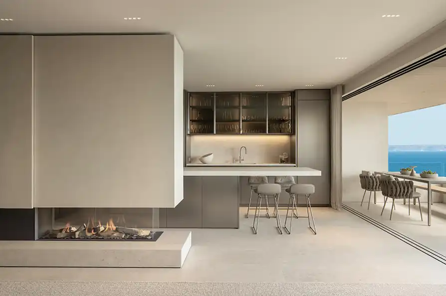 Modern kitchen with beige and gray tones featuring a minimalist gas fireplace on the left, a sleek bar counter with two gray bar stools in the center, glass cabinets with dishware above the counter, and a large open sliding glass door leading to an outdoor dining area with a view of the ocean.