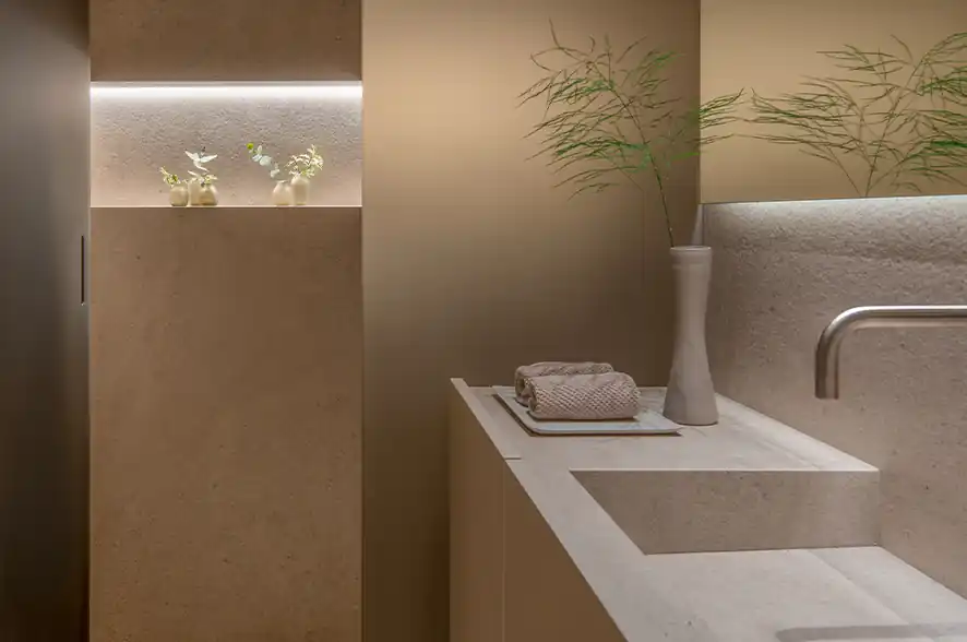 Minimalist bathroom vanity with modern beige stone sink, a sleek metal faucet, a rolled towel on a tray, and a tall white vase with delicate green branches, next to a wall niche with small vases and plants, all softly illuminated with warm lighting.