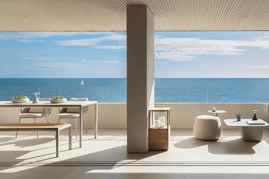 Modern outdoor patio with minimalist furniture including a dining table with chairs on the left and low tables with cushioned stools on the right, overlooking a vast blue ocean with a small white sailboat in the distance under a partly cloudy sky.