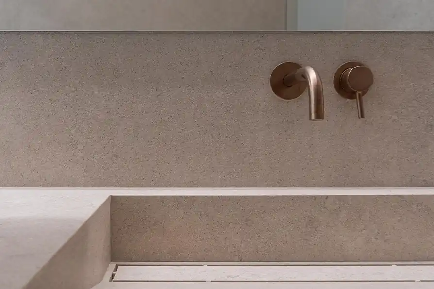 Minimalist bathroom sink with beige stone countertop and wall, featuring a modern brushed bronze wall-mounted faucet and lever handle.