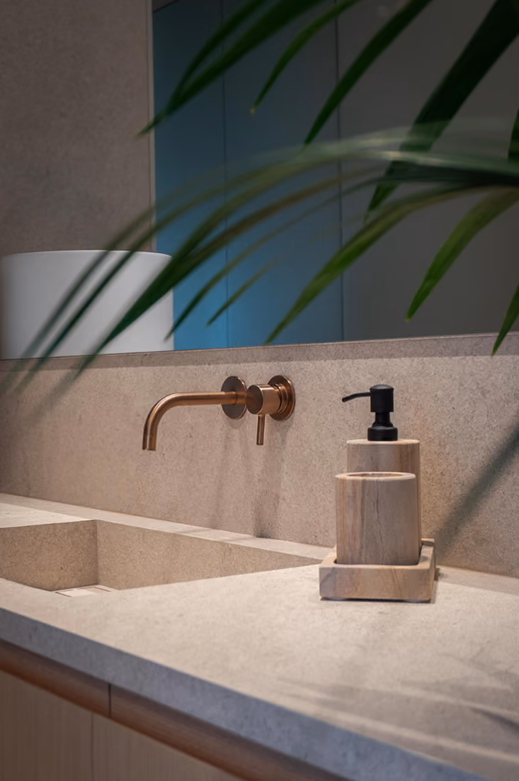 Minimalist bathroom sink with a beige stone countertop, an inset rectangular basin, a wall-mounted copper faucet, and a wooden soap dispenser and tumbler set on a matching tray, partially obscured by green plant leaves.