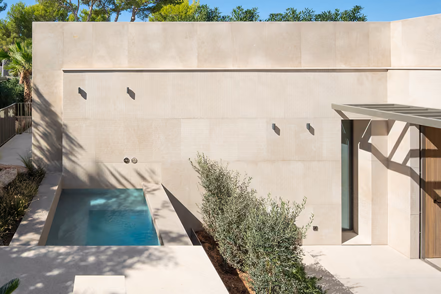 Modern outdoor patio with beige stone walls and flooring, a small rectangular pool with clear blue water, green shrubbery planting along the pool edge, and tall trees in the background under a clear blue sky.