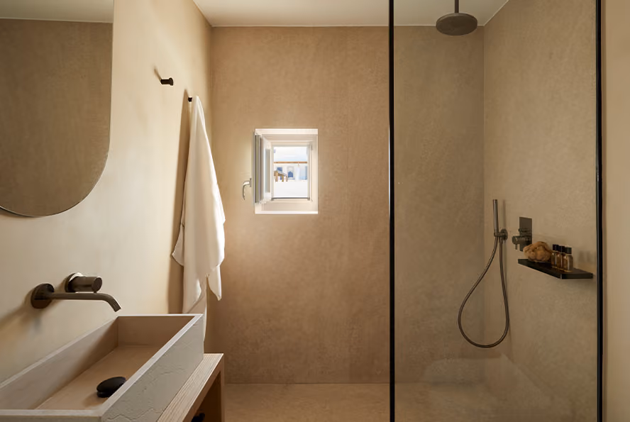 Minimalist bathroom with beige walls, a rectangular sink with black soap, a round mirror, a hanging white towel, a small window letting in natural light, and a glass-enclosed shower area with a rain showerhead, handheld shower wand, and toiletries on a wall shelf.