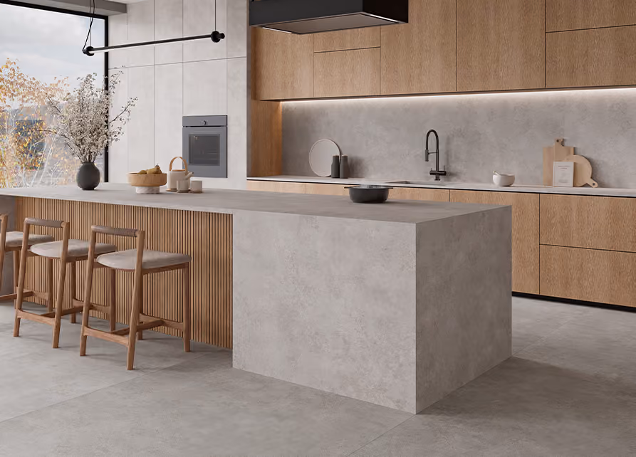 Modern kitchen with a large concrete island featuring three wooden chairs, wood cabinets, a built-in oven, a black faucet, decorative vases, cutting boards, and a large window with a view of autumn trees.