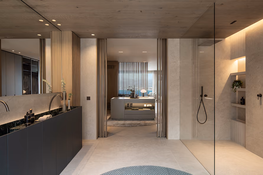 Modern spacious bathroom with a double black vanity and marble countertop to the left, a large walk-in glass shower to the right, and a view through sliding glass doors into a bedroom with a central dresser and sheer curtains revealing a scenic outdoor view.