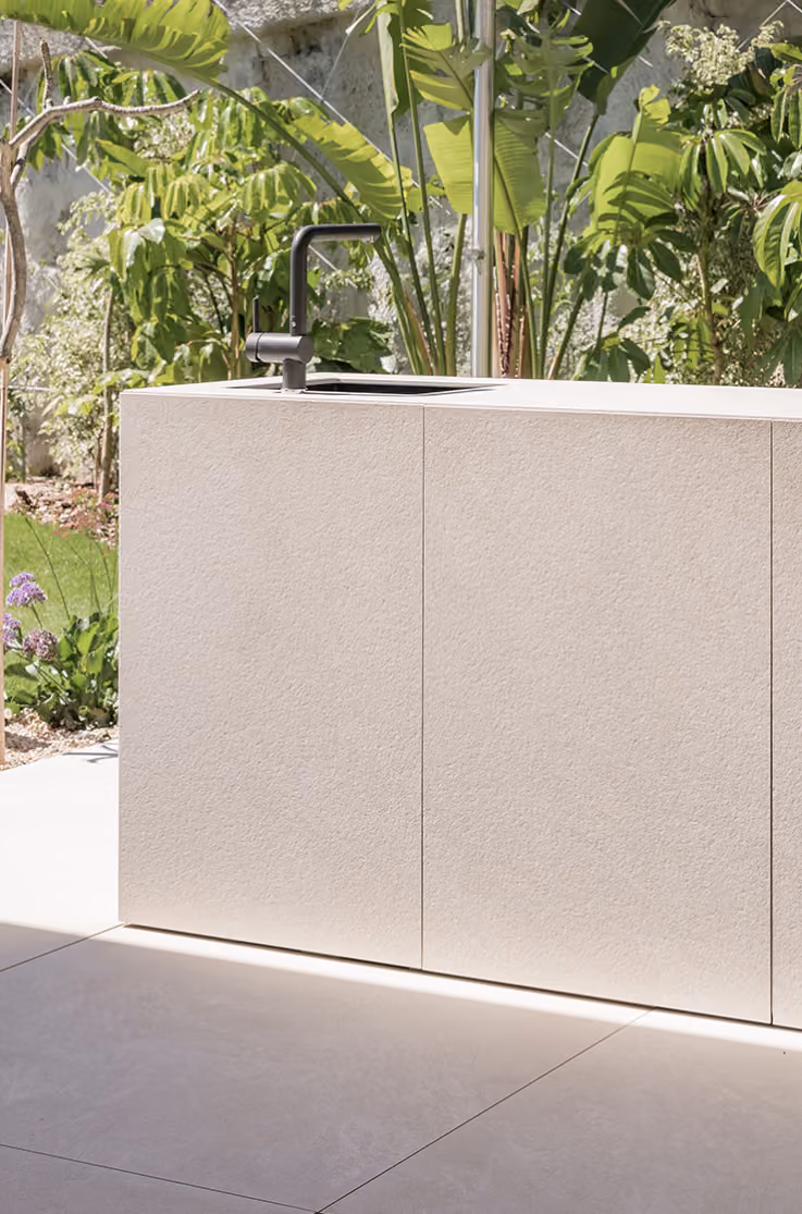 Minimalist outdoor kitchen island with a light beige textured surface, featuring a black faucet and sink, set against lush green tropical plants.